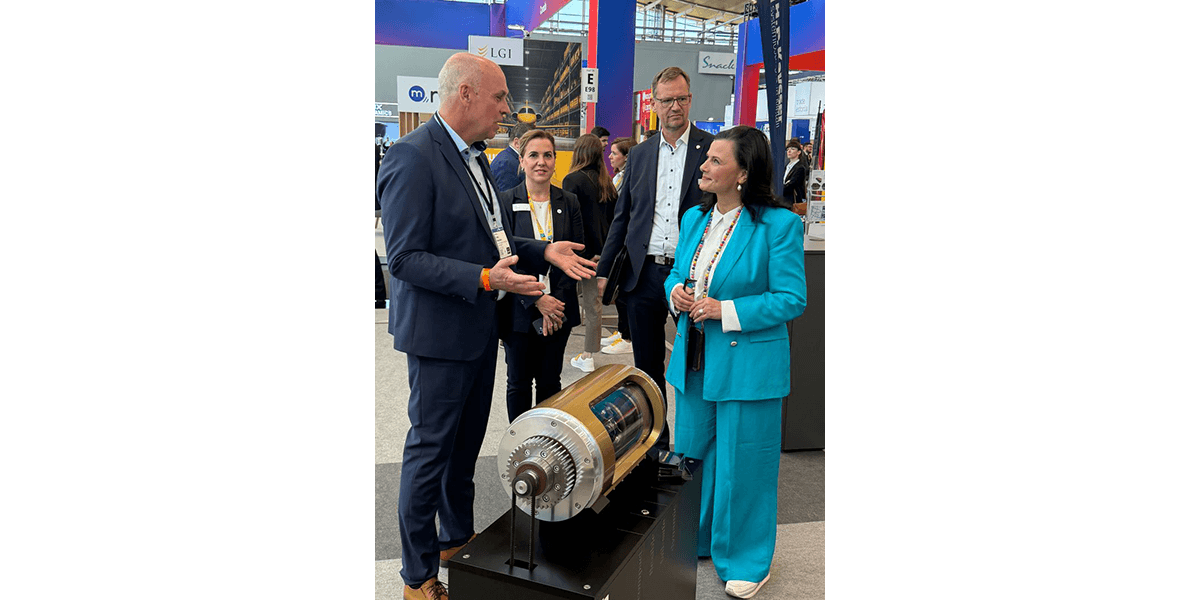 Sytuacja na targach przemysłowych: kilka osób rozmawiających przy stoisku, na pierwszym planie wielkoformatowy model przekrojowy mechanicznego elementu napędowego umieszczony na podwyższeniu.