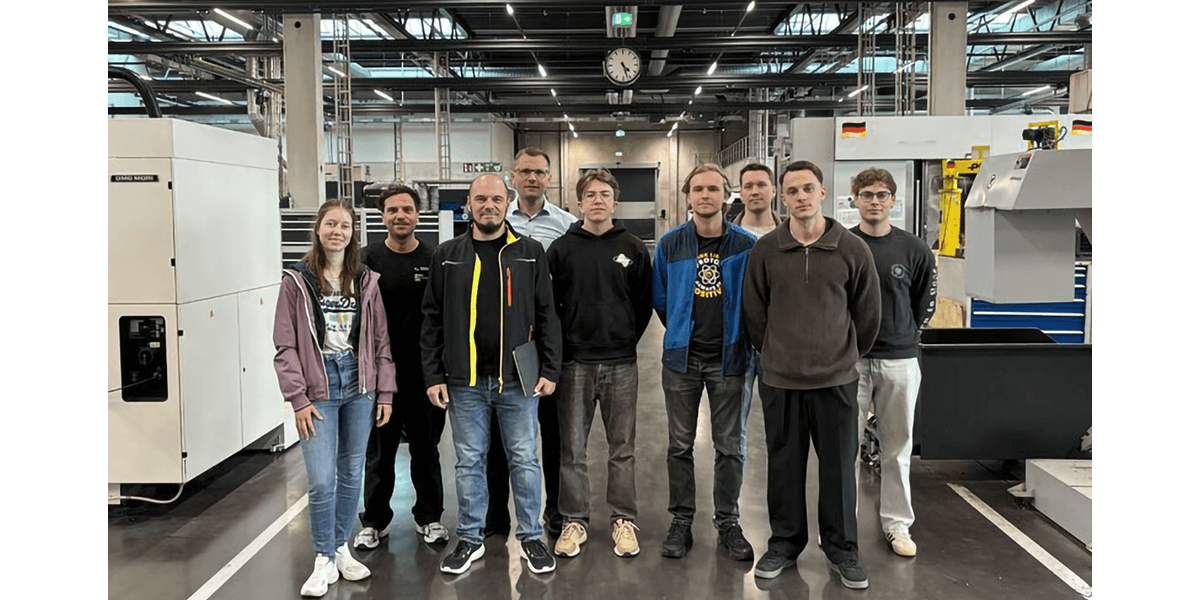 Gruppe von neun Personen steht in einer modernen Produktionshalle zwischen großen CNC‑Maschinen und Industrieanlagen.